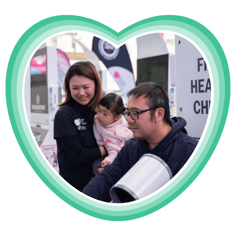 A woman holding a young child smiles beside a man taking a free health check at a self-serve Shane Warne Legacy station