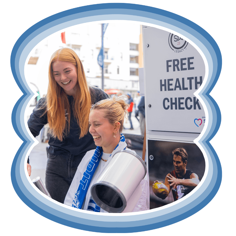 Two women laughing together at a free Shane Warne Legacy health check station on a game day