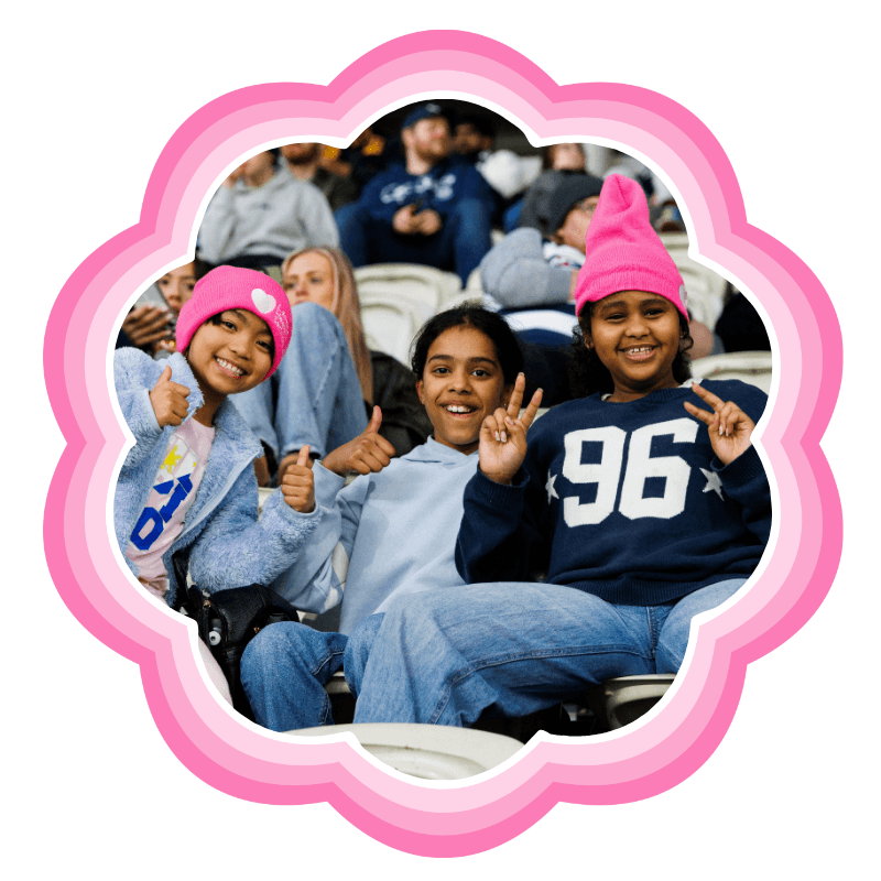 Three young North Melbourne supporters in beanies smiling and giving a thumbs up from the stands